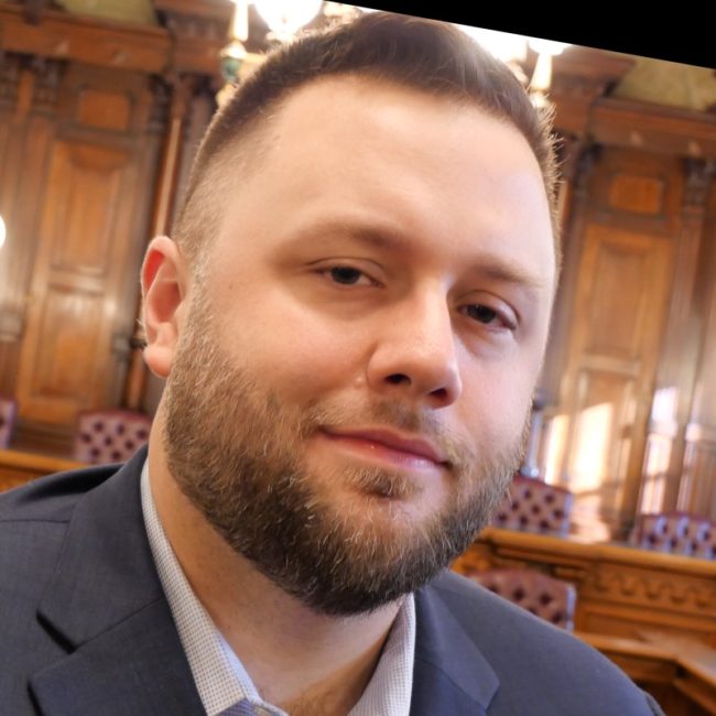 1681339611663 Image of Shane Trejo in front of House chamber. He is wearing a navy suit jacket. He has a beard and his hair has a skin fade across the sides.