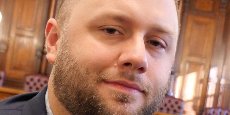 1681339611663 Image of Shane Trejo in front of House chamber. He is wearing a navy suit jacket. He has a beard and his hair has a skin fade across the sides.