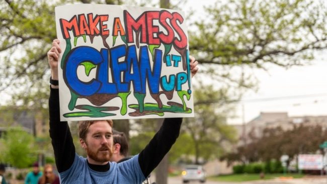 Image of a bearded white man holding a sign in front of a tree that reads "Make a mess clean it up." The font is orange, red and blue and has green and brown sludge oozing out.