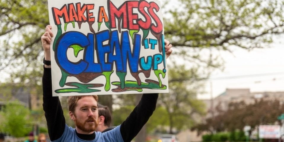 Image of a bearded white man holding a sign in front of a tree that reads "Make a mess clean it up." The font is orange, red and blue and has green and brown sludge oozing out.
